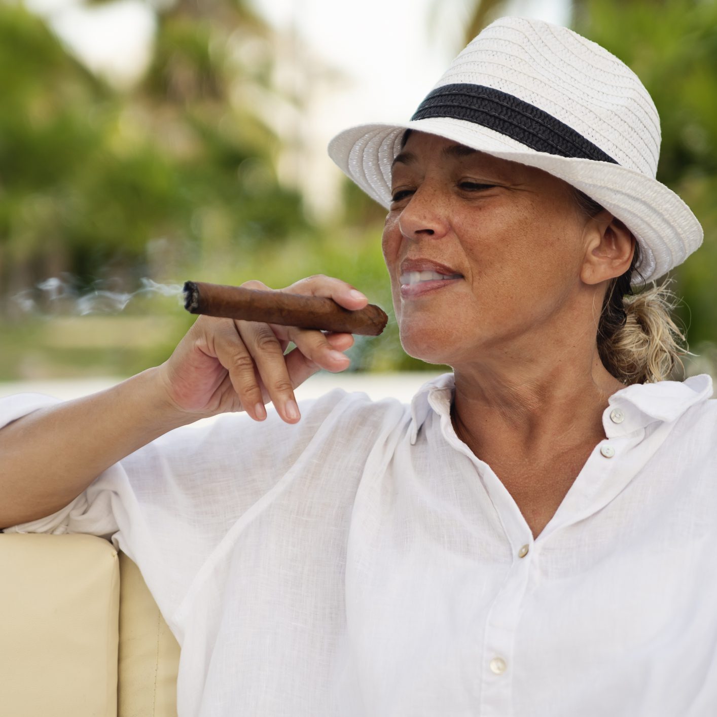 Beautiful 50+ woman smoking a cigar in a tropical setting. She is wearing a white straw hat, and a long white shirt. She is tanned and relaxed, enjoying the moment. Horizontal waist up outdoors shot with copy space. This was taken in Cayo Largo, Cuba.